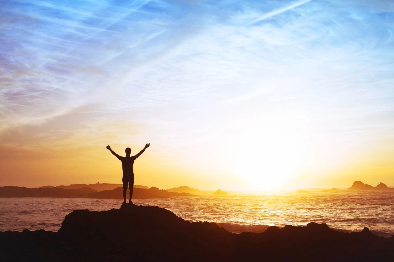 Person celebrating on a hill during sunrise by the ocean.